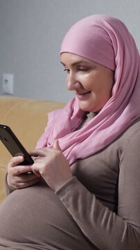 Smiling pregnant muslim woman in a traditional pink hijab enjoying her leisure time at home, scrolling through social media on her smartphone and resting comfortably on the couch