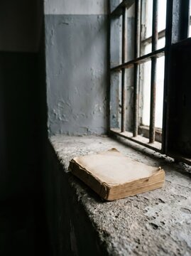 Old worn book lying on concrete windowsill inside prison cell with rusted metal bars. Concept of education, hope and isolation during confinement. Knowledge as freedom behind bars