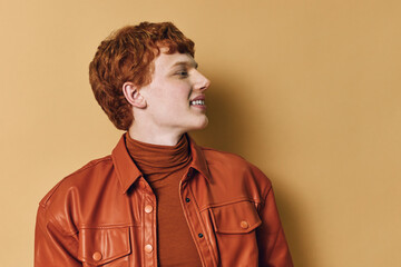 Obraz premium Young man with red hair wearing a brown leather jacket and turtleneck looking sideways smiling against a beige background in studio portrait.
