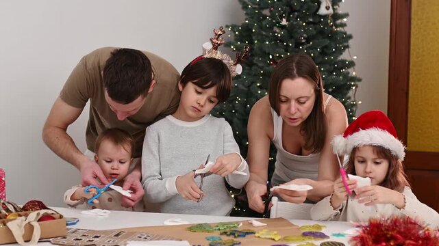 Family celebrates Christmas together as parents and children craft festive paper decorations at table beside glowing Christmas tree, enjoying creative holiday time at home