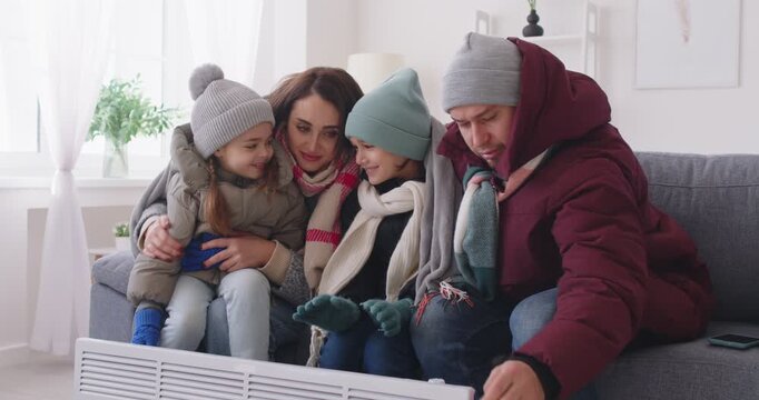 Freezing parents and children, dressed in winter outerwear, gather near a heater, sitting on sofa at home to escape the freezing weather. Warmth, family unity, and relief from the cold.