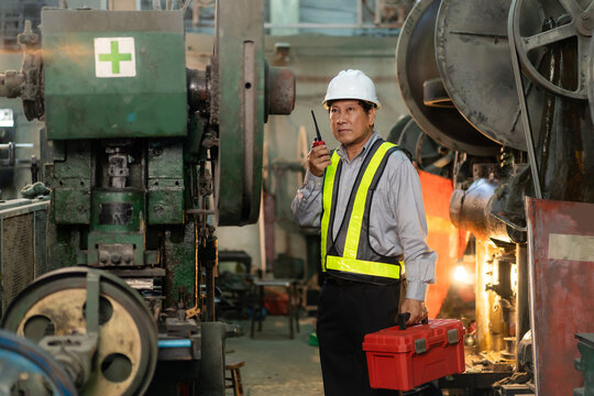 Supervisor wearing hardhat holding walkie talkie and toolbox to inspect machinery safety in factory.