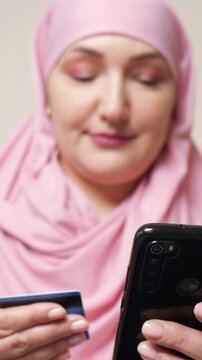 Young muslim woman in a pink hijab with beautiful makeup making an online purchase using her credit card and smartphone, embodying the concept of modern electronic commerce and banking