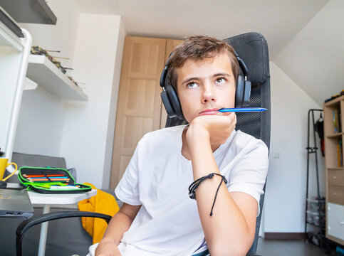 Teenage boy doing homework with headphone at home. Student feeling boring, uninteresting, concentration struggle. Caucasian male model resting his head on his hand while looking up and dreaming.   
