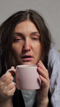 Young unwell woman with flu symptoms, looking tired and sick, wrapped in a blanket, drinking hot tea from a pink mug for relief, experiencing fever, runny nose, and coughing at home