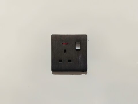 ​Modern black UK wall power socket with switch and red indicator light on a plain white wall background.