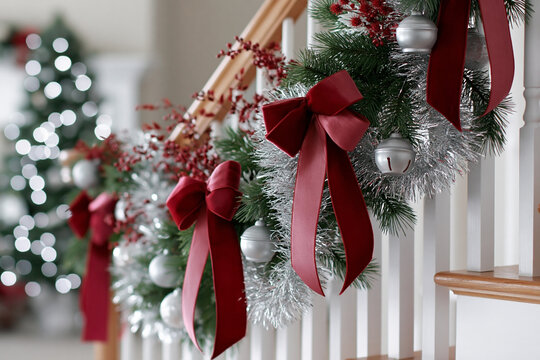 Christmas staircase decorated with evergreen garland, red bows, silver tinsel and bells with blurred festive tree lights in background