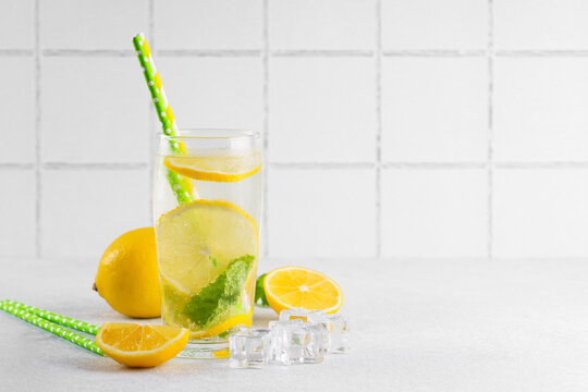 Refreshing homemade lemonade served in clear glass with fresh lemon slices, mint leaves, ice cubes and paper straws on a light kitchen counter against a white tile background with copy space.