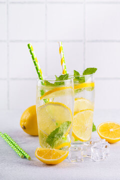 Close up of two clear glasses with refreshing homemade lemonade with fresh lemon slices, mint leaves, ice cubes and paper straws on a light kitchen counter against white tile background