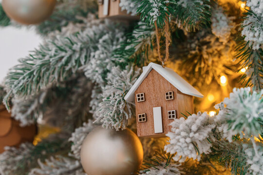 Cozy Christmas tree snow covered branch decorated with wooden house ornament, warm lights garland and golden baubles, creating festive winter atmosphere. Festive aesthetic atmosphere, holiday mood