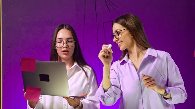 Close up of colleagues planning strategy on purple glass board. One holds laptop with sticky notes while other draws diagram overhead.