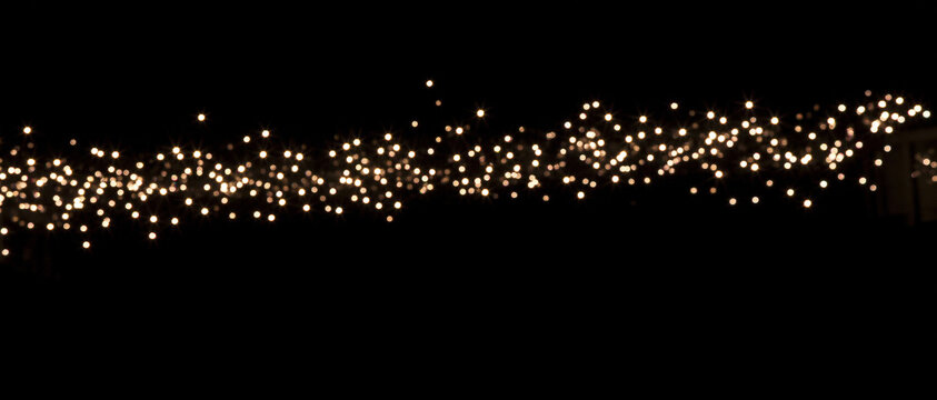 Horizontal border of glowing fairy lights against black background. Sparkling bokeh garland with warm white festive illumination. Abstract holiday banner with copy space