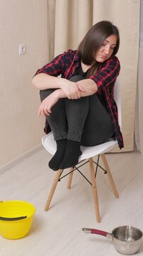 Frustrated brunette woman sitting on a chair, looking up with despair as water from a ceiling leak drips into a bucket and a pan, showing the stress of a flooded apartment emergency