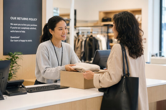 Retail clerk hands a boxed item back to a customer at a store counter while smiling and speaking by the register near a returns policy sign.
