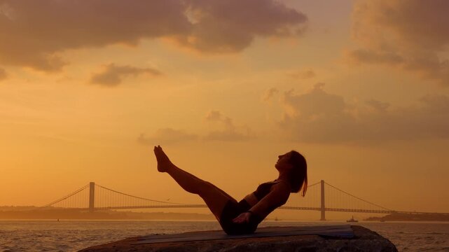 Woman performing boat pose exercise at twilight near New York bay.. Full silhouette of female balancing in v shape position. Warm orange sky, water and distant bridge in background.