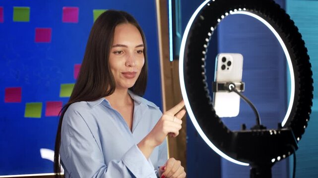 Content creator gesturing during live stream recording session. Female influencer talking and pointing finger toward smartphone camera inside broadcast studio.
