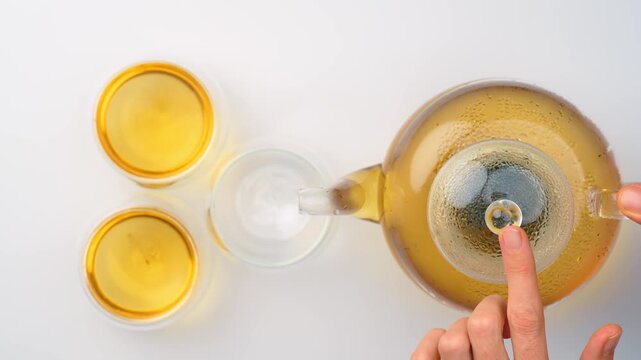 Hand holding glass teapot lid beside three cups with melissa tea. Top view flat lay of yellow drink on white background.
