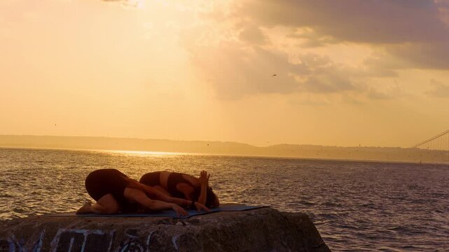 Women resting in child pose on concrete block by ocean. Two practitioners bow forward on mats with golden sky overhead near Verrazzano Narrows Bridge, New York.