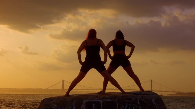 Two women standing in wide stance yoga posture at sunset in New York. Silhouetted female duo on concrete block facing Verrazzano Narrows Bridge over water. Golden clouds above.