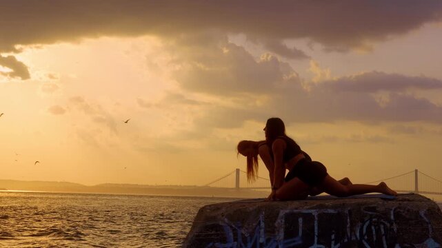Women practicing pigeon pose on stone block near sea. Two female athletes hold low lunge position on waterfront pier during golden hour in New York.