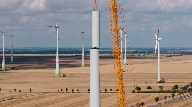 Aerial drone view wind turbine installation across harvested farmland, with cranes lifting tower sections. Renewable energy construction expands wind farm infrastructure under dramatic summer clouds