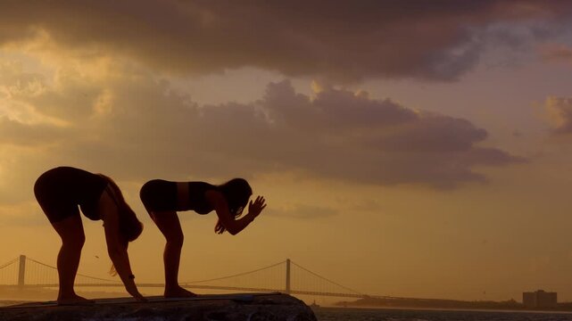 Women leaning down in yoga pose near bay. Two female silhouettes execute standing forward bend on pier with Verrazzano Narrows Bridge behind.