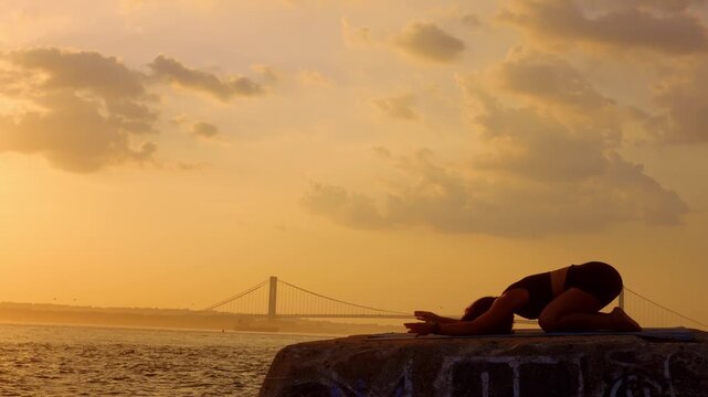 Woman practicing child pose on mat during sunset near New York bridge. Profile silhouette of female figure resting on concrete block near water. Warm evening lighting and clouds above.