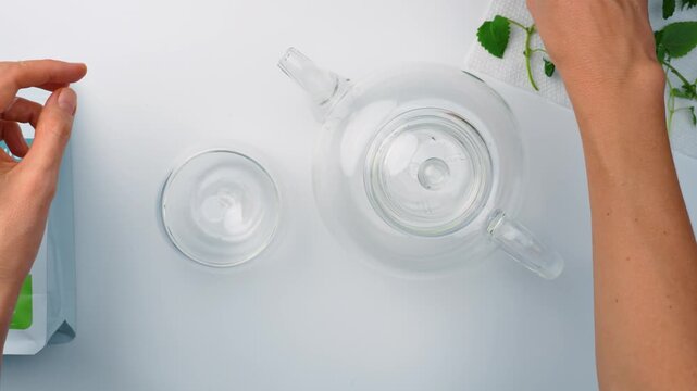 Glass teapot and cup with hand on white surface. Overhead angle of transparent tea set and fresh green herbs with arm reaching from side.