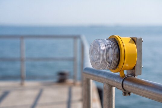 A close-up of a bright yellow industrial warning light fixture mounted on a stainless steel railing at a concrete pier, with the blurred blue sea in the background.