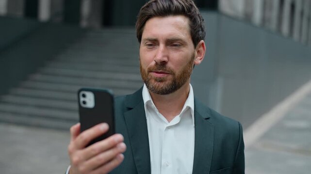 Happy smiling Caucasian middle-aged business man holding phone talking video call mobile virtual chat in city near office building. Cheerful mature businessman male employer talk smartphone conference