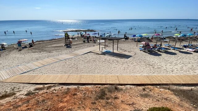 Playa time lapse, arena y mar con ba&ntilde;istas, pasarela de madera, tumbona hamaca y socorrista vacaciones de verano y sol, horizonte brillo reflejo en agua azul mar&iacute;timo velocidad v&iacute;deo r&aacute;pido acelerado