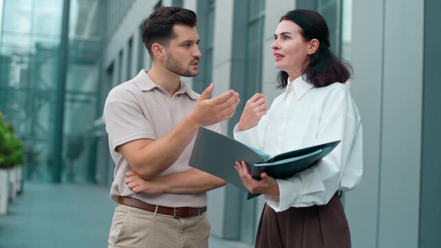 Two troubled Caucasian business colleagues talking quarreling looking in documents in city. Intern man talk arguing with angry woman annoyed with report mistake businessman argue with businesswoman