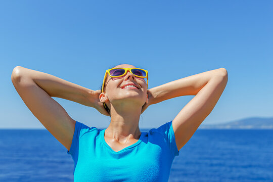 A happy woman with yellow sunglasses relaxes in the sun against a clear blue sky and ocean backdrop.