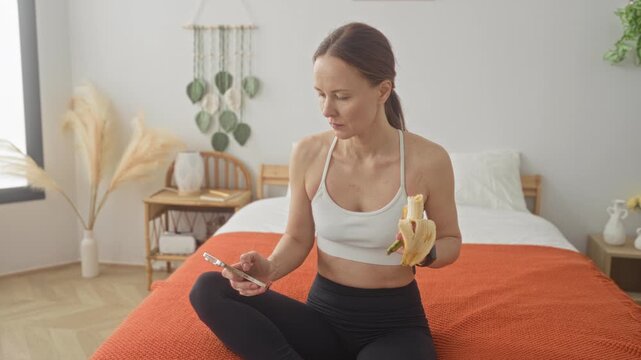 Young woman in sports bra holding a peeled banana and checking a smartphone while seated on bed in studio; post workout snack calm.