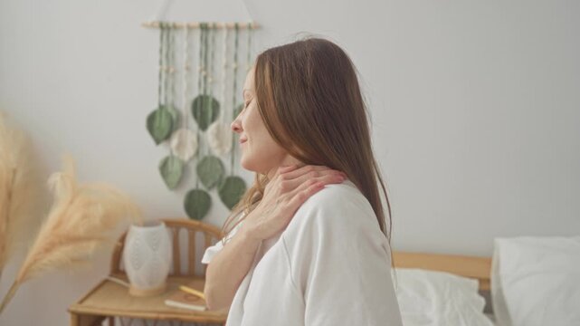 Young woman massaging shoulder in bedroom beside bed and macrame wall hanging, eyes closed in morning light; self care relief.