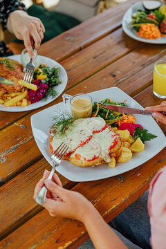 Diners enjoy plated meals with fries, salad, vegetables, sauce, and colorful drinks at a rustic wooden outdoor table in a casual restaurant setting