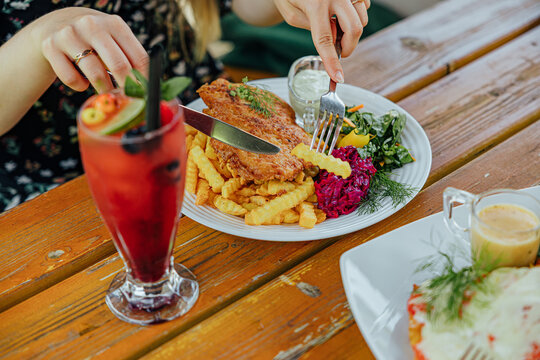 Diner cuts into a crispy fried cutlet served with crinkle fries, beet salad, greens, dill, and creamy sauce beside a chilled red berry drink on a wooden table