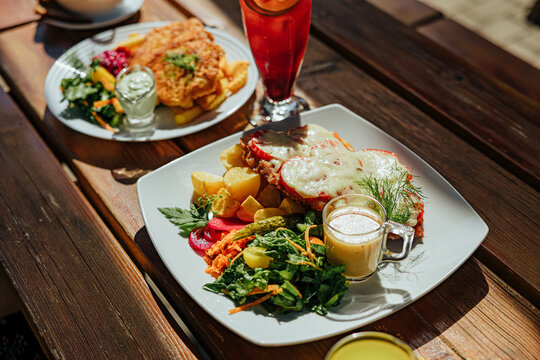 Plated restaurant meal with cheese-topped meat, roasted potatoes, fresh salad, pickled vegetables, creamy sauce, and a red fruit drink on a sunlit wooden table