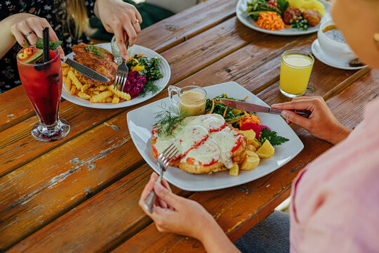 Diners enjoy plated meals with fries, salad, vegetables, sauce, and colorful drinks at a rustic wooden outdoor table in a casual restaurant setting