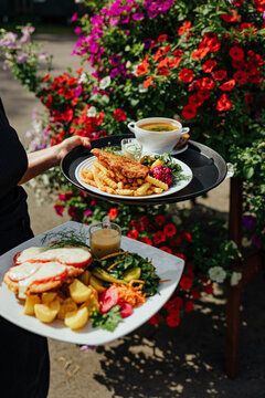 Restaurant server carries two plated meals with fries, potatoes, salad, sauce, and soup through a bright outdoor garden filled with colorful flowers