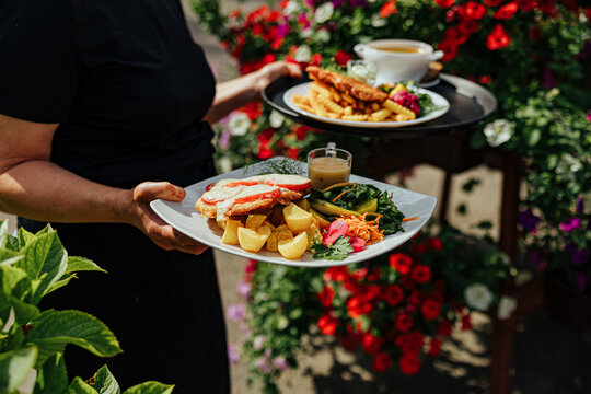 Restaurant server carries two plated meals with fries, potatoes, salad, sauce, and soup through a bright outdoor garden filled with colorful flowers