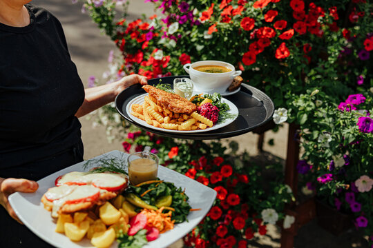 Restaurant server carries two plated meals with fries, potatoes, salad, sauce, and soup through a bright outdoor garden filled with colorful flowers