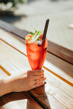 Close-up of a hand holding a chilled red fruit drink with ice, berries, mint, and straws on a sunlit wooden table, with soft background copy space