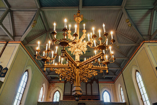 Elaborate golden chandelier with glowing candle-style lights beneath a wooden vaulted ceiling, framed by arched church windows and a pipe organ