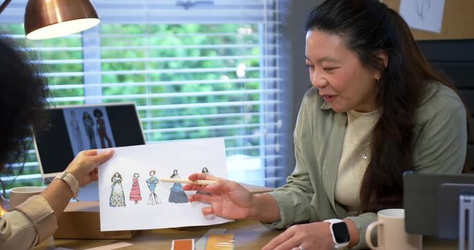 Asian woman pointing pen peer holding five sketches reviewing colors at desk by window, copy space