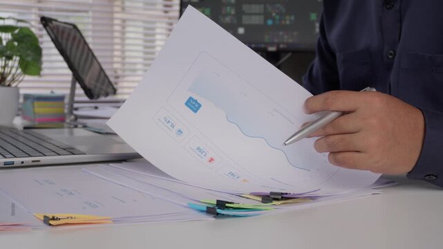 Business professional analyzing financial charts and performance data in printed reports while turning document pages, reviewing statistics and information for strategic planning and decision making
