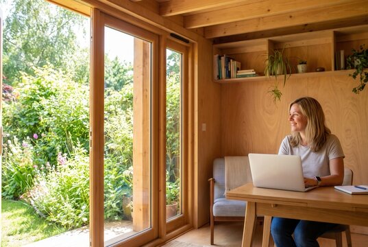 Female working on laptop at wooden desk in bright home office with large windows overlooking a lush garden filled with greenery and flowers