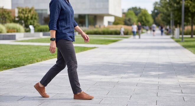 Side view of person walking on paved walkway in modern urban park campus wearing fitness tracker smartband in blue casual shirt and brown leather sneakers daylight