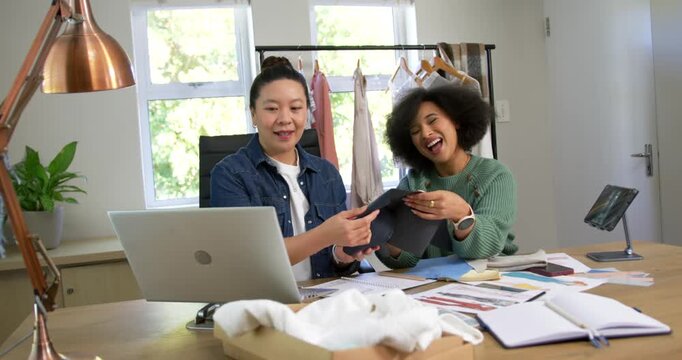 African American, Asian women exchanging, inspecting dark fabric at studio using laptop for colors