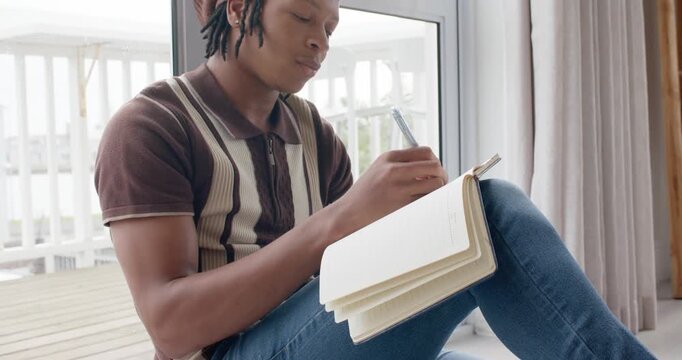 African man sitting by glass door, journaling in open notebook, pen moving as daylight inspiring